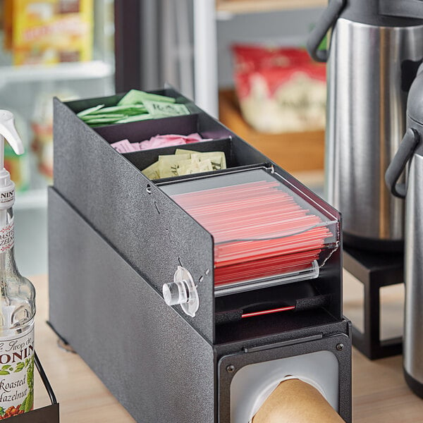 A black KleanTake dispenser holding unwrapped stirrer straws in a plastic container.