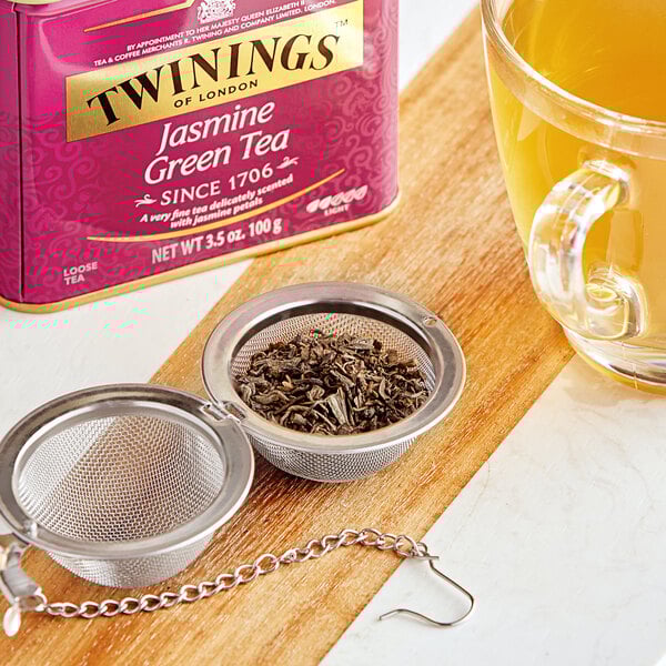 A strainer full of Twinings Jasmine Green loose leaves over a cup of orange liquid.