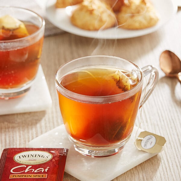 A glass cup of Twinings Pumpkin Spice Chai Tea with a tea bag next to it on a saucer.