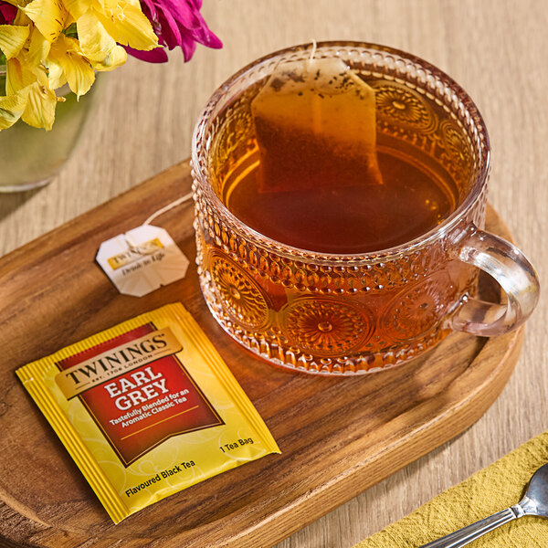 A glass cup of Twinings Earl Grey tea with a tea bag on a tray next to a yellow flower.
