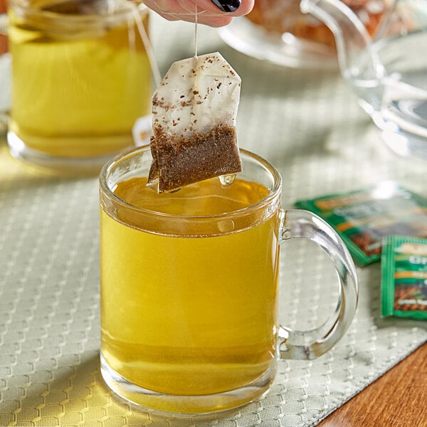 A hand using a Twinings Green Decaffeinated Tea bag to pour tea into a glass cup.