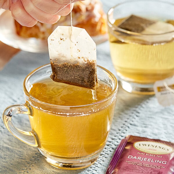 A hand holding a Twinings Darjeeling tea bag over a glass cup of tea.