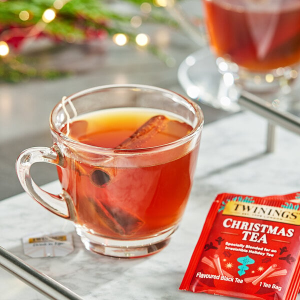 A glass cup of Twinings Christmas tea with cinnamon sticks and a red packet of Twinings tea on the table.