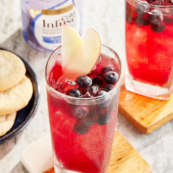 A glass of red Twinings Blueberry & Apple cold water with blueberries and apple slices on a table with a cookie on a plate.