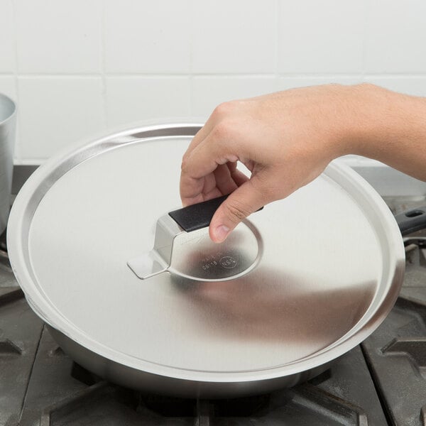 A hand using a Vollrath Tribute lid with a Torogard handle to cover a pan on a stove.