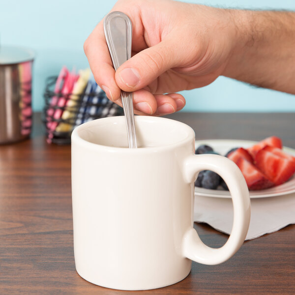 A hand holding a spoon in a white Tuxton china mug.