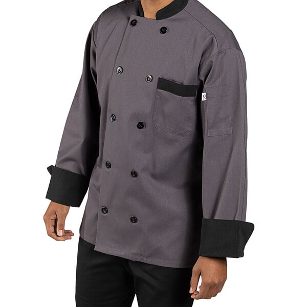 A person wearing a Uncommon Chef Newport long sleeve chef coat with black trim in a professional kitchen.