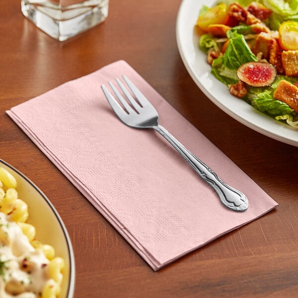 A fork on a pink Hoffmaster dinner napkin next to a plate of salad.