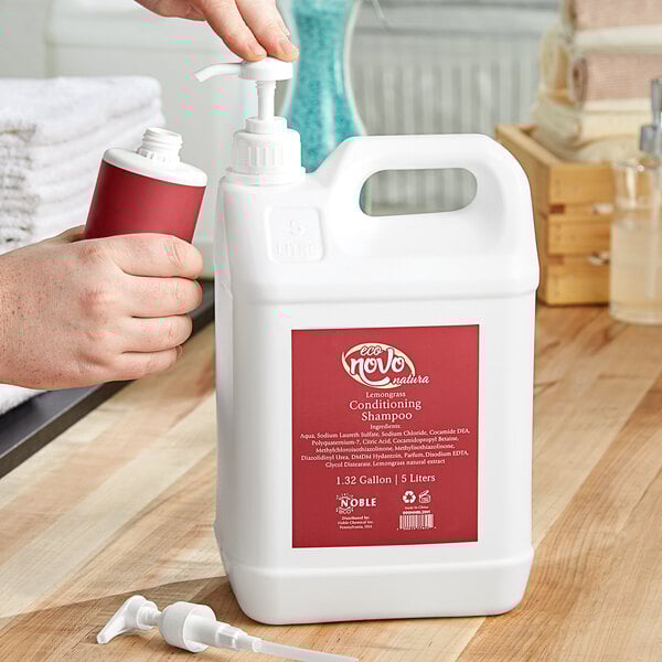 A person using a pump dispenser to hold a white plastic bottle of Noble Eco Novo Natura conditioning shampoo with a red label