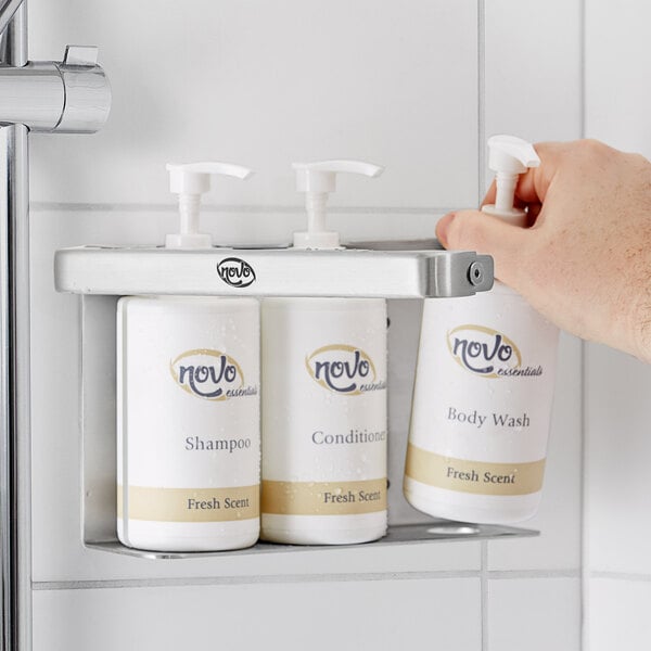 A hand holding a white plastic pump dispenser with Novo by Noble Chemical Triple Bottle Bracket for hotel amenities on a shelf.