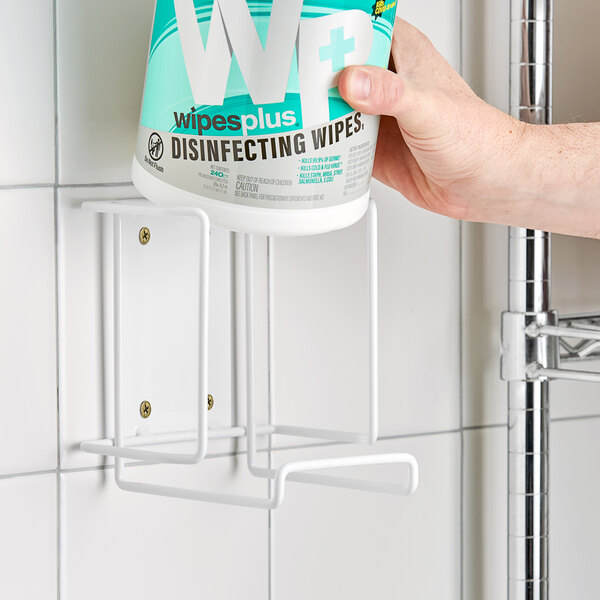 A hand holding a WipesPlus wall bracket with a container of wipes on a wall.