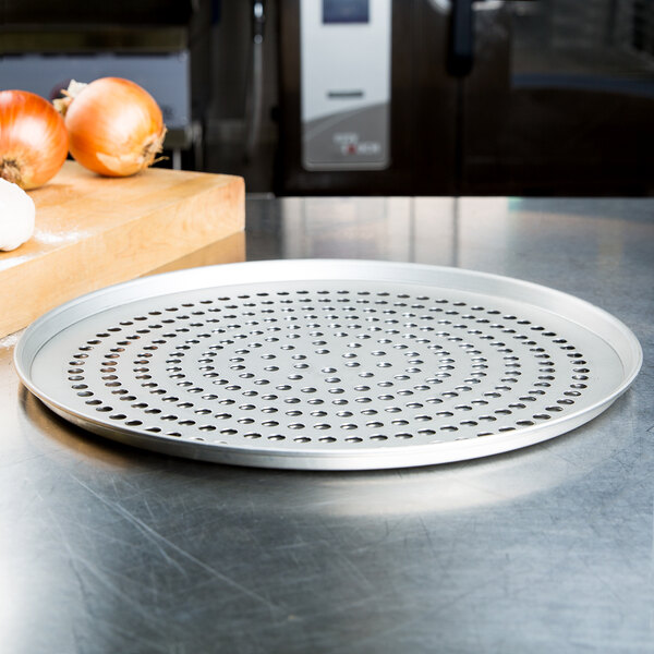 An American Metalcraft tapered pizza pan with holes on a white background next to onions.