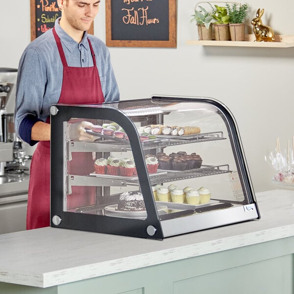 A man in a red apron standing behind a Avantco countertop bakery display case filled with cupcakes.