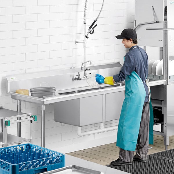 A person in a blue apron washing dishes in a Regency 3-compartment dish table.