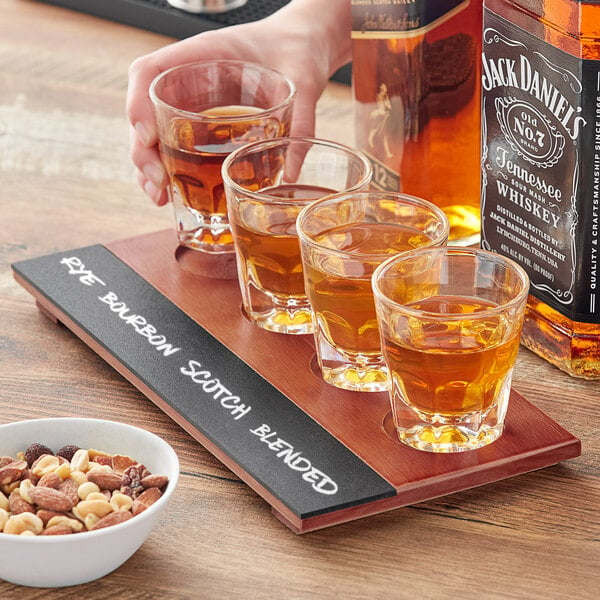 An Acopa flight tray with espresso glasses filled with amber liquid on a table with a bowl of nuts.