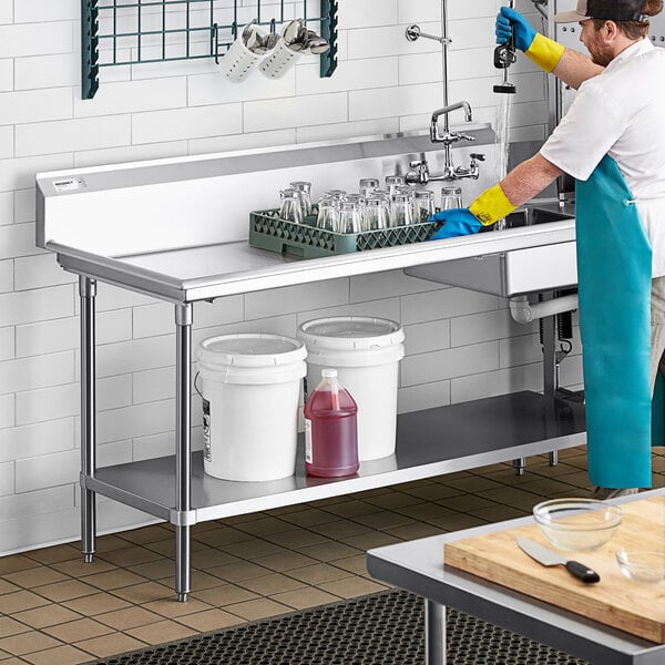 A stainless steel dish table with an undershelf, holding two white buckets and a red container, is shown in a commercial kitchen setting.