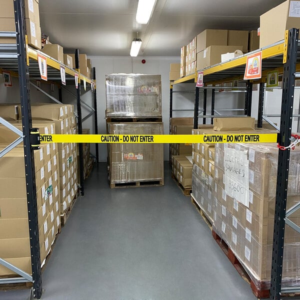 A yellow Banner Stakes wall mount barrier tape with "Caution - Do Not Enter" on shelves in a warehouse.