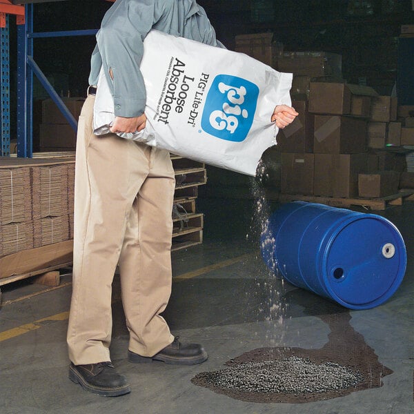 A person pouring New Pig Lite-Dri loose absorbent from a bag onto a liquid spill near a blue drum in a warehouse.