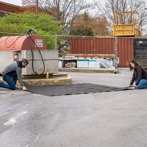 A large black absorbent ground tarp being laid out by two people near an industrial fuel tank.