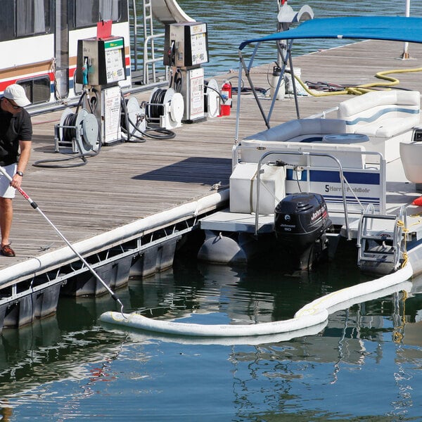 A white absorbent boom floating on water near a docked boat, designed for containing oil-based liquid spills.