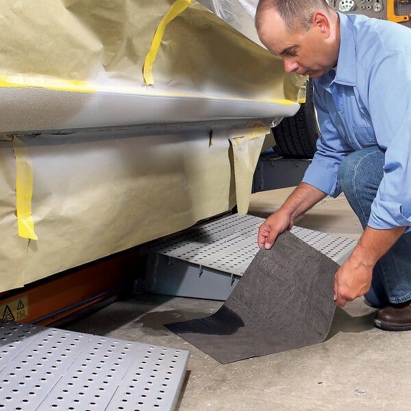 A person placing a black absorbent mat pad on the floor near a vehicle covered in masking paper.