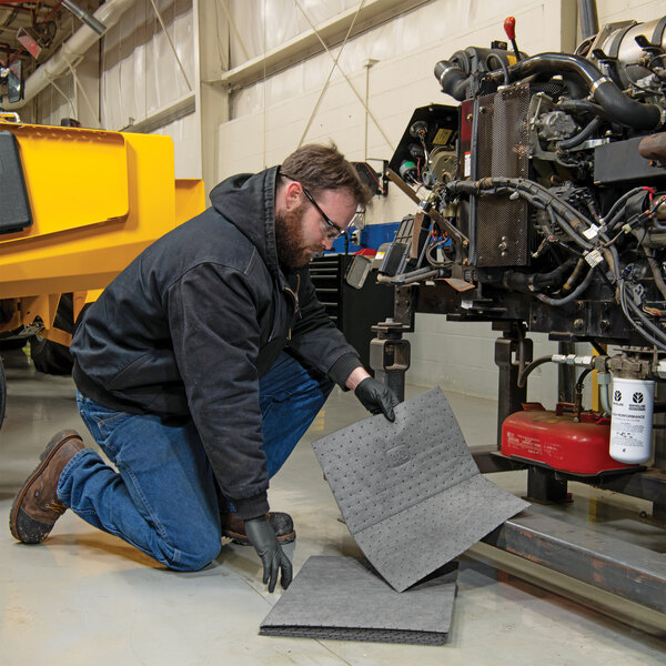 A person placing a gray absorbent mat pad on the floor near machinery in a workshop setting.
