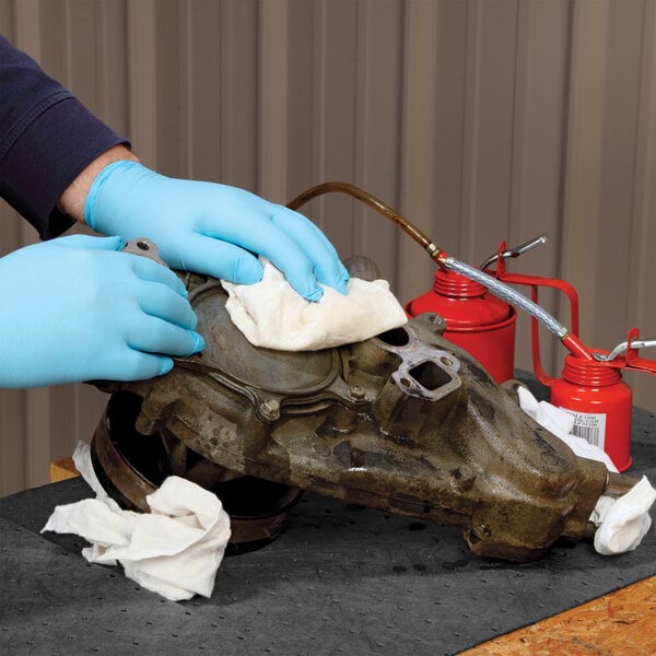 A person wearing blue gloves uses a white cloth rag to clean a greasy metal part on a workbench.
