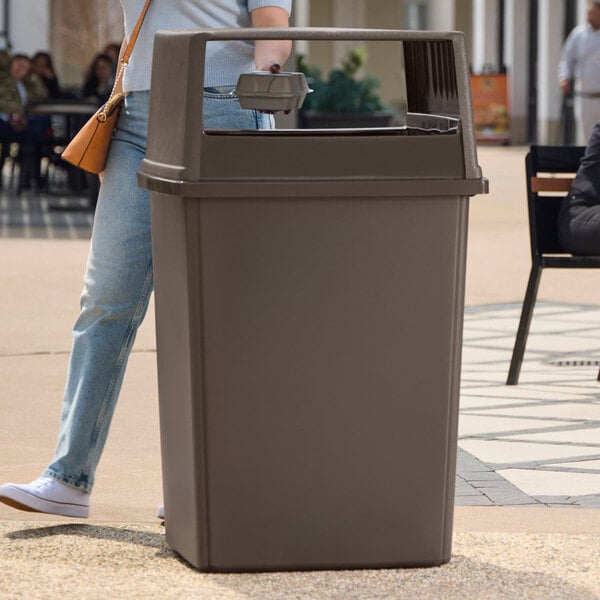 A large, brown, square trash can with an open top is placed outdoors near a person walking by.