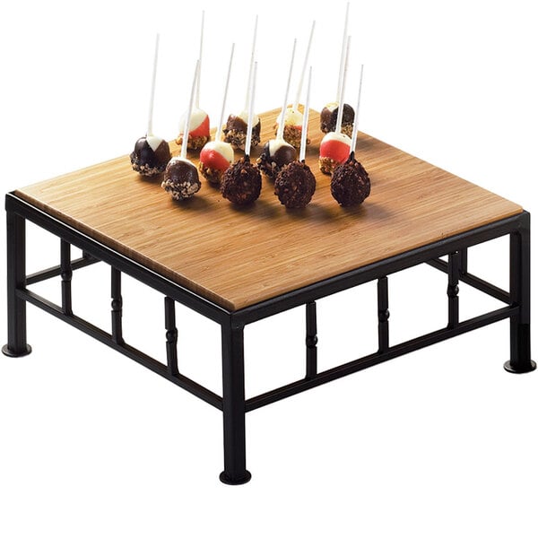 A Cal-Mil iron black square riser with a bamboo top on a wooden table with cake pops on skewers.