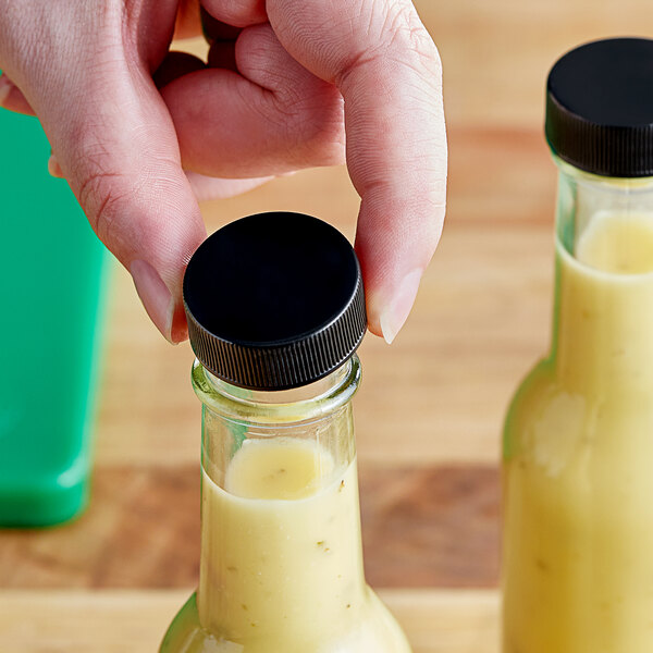 A hand holding a bottle of sauce with a black ribbed plastic lid.