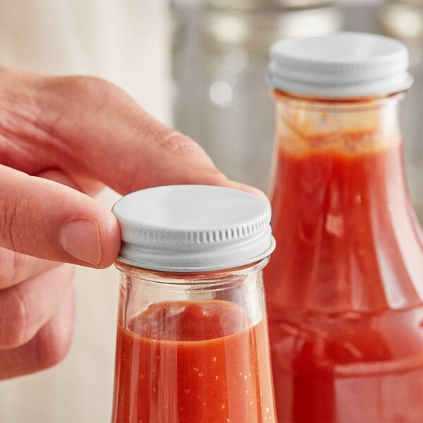 A hand holding a bottle of red sauce with a 38/400 White Metal Lid.