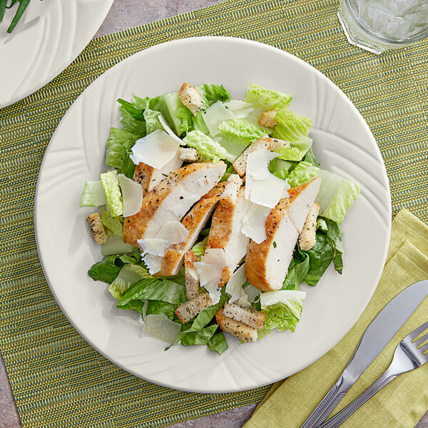 An Acopa Swell ivory stoneware plate with a wide rim and embossed design holding a chicken salad plate with a fork and knife.