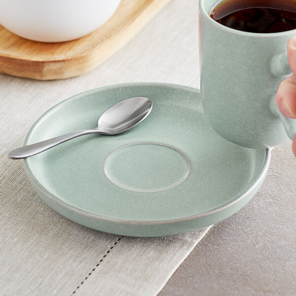 A hand holding a cup of coffee on a Acopa Harbor Blue saucer with a spoon.