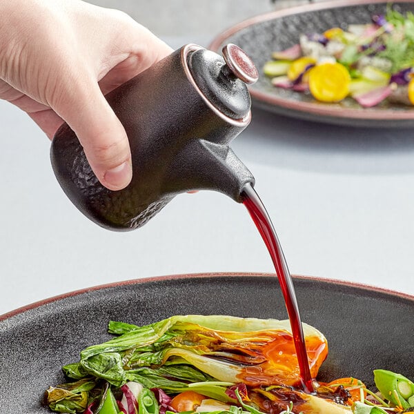 A hand pouring soy sauce from a black stoneware pourer onto a bowl of vegetables.