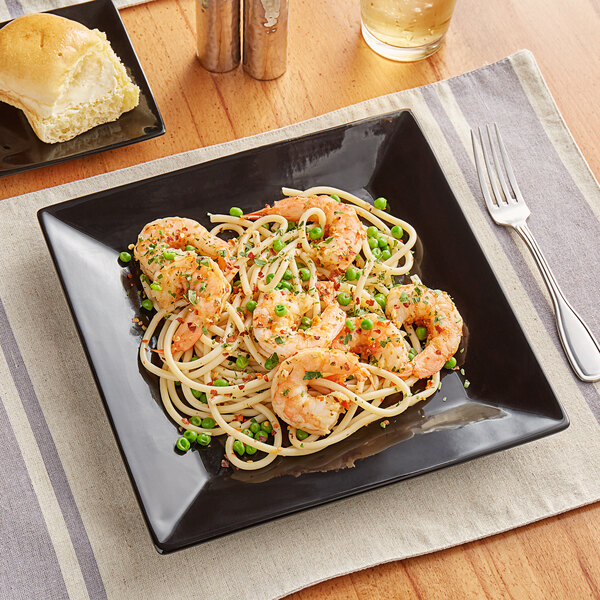 A glossy black square Acopa stoneware plate with spaghetti, shrimp, and peas on it.