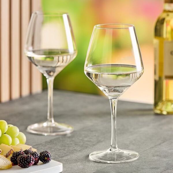 Two Acopa Silhouette wine glasses filled with white wine on a table with grapes and cheese.