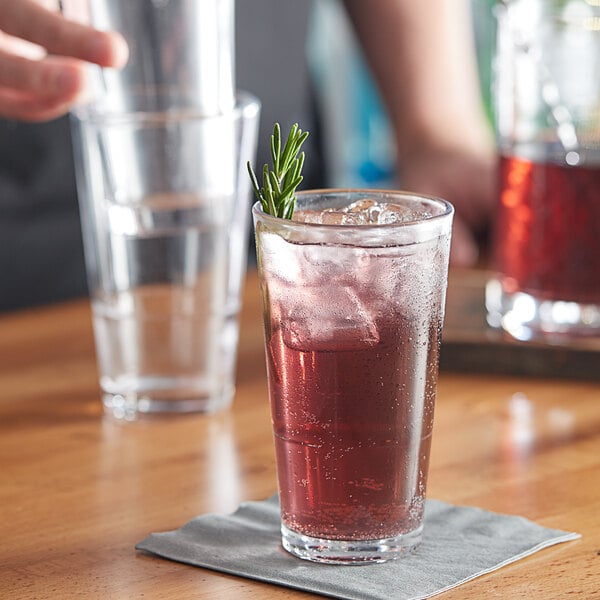 A person holding an Acopa Select Highball Glass filled with red liquid on a table with a napkin and a sprig of rosemary.