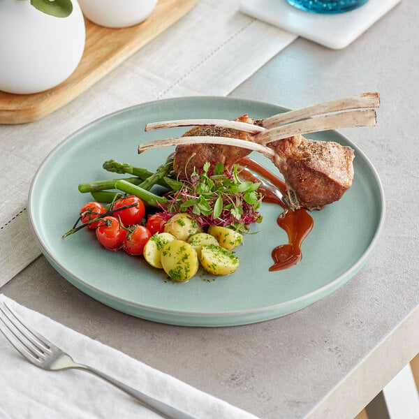 An Acopa Pangea Harbor Blue coupe plate with ribs, meat, and vegetables on it on a table.