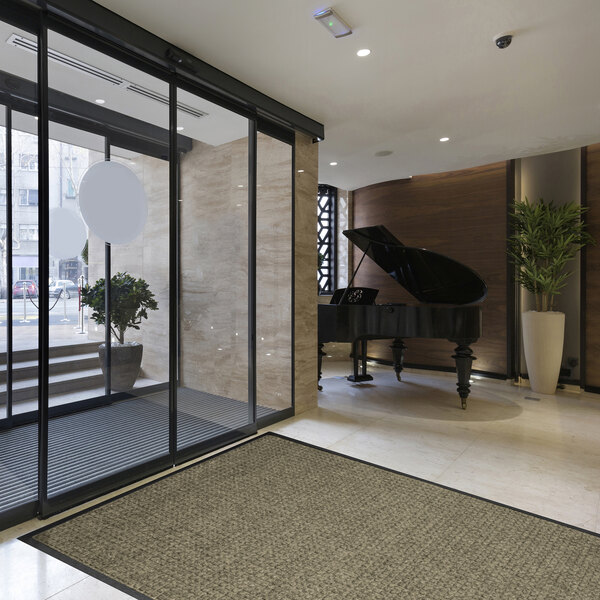 A modern lobby area with a large gray floor mat near glass entrance doors, a grand piano, and potted plants.