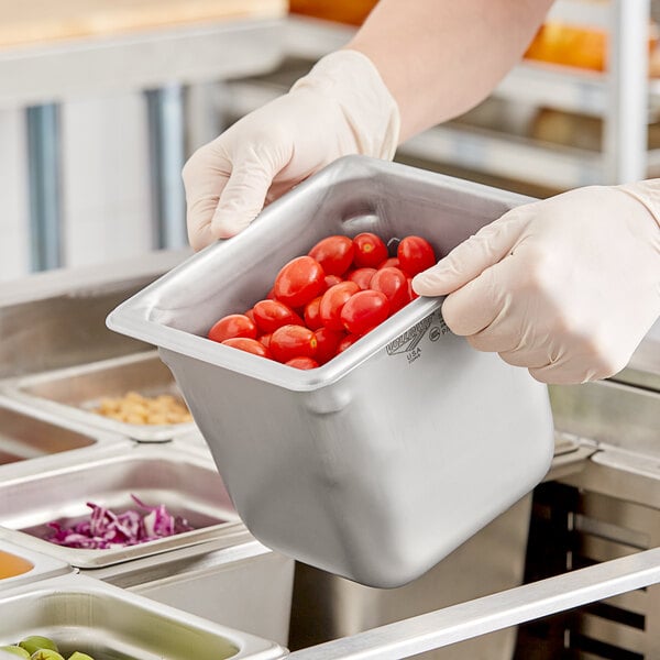 A person in gloves holding a Vollrath stainless steel steam table pan full of tomatoes on a counter.