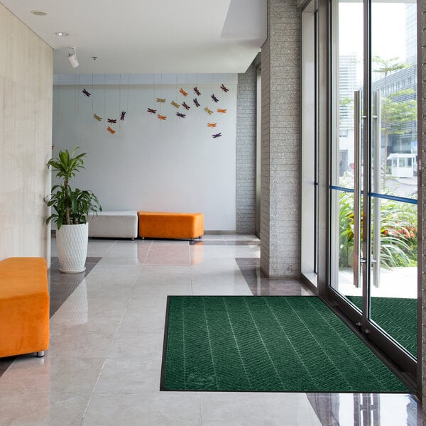 A green rectangular floor mat with a classic border is placed near the glass entrance doors of a modern lobby with tiled floors, orange and white seating, and wall decorations.