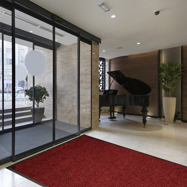 A modern lobby area with a large red mat on the floor, a grand piano in the background, and glass doors leading outside.