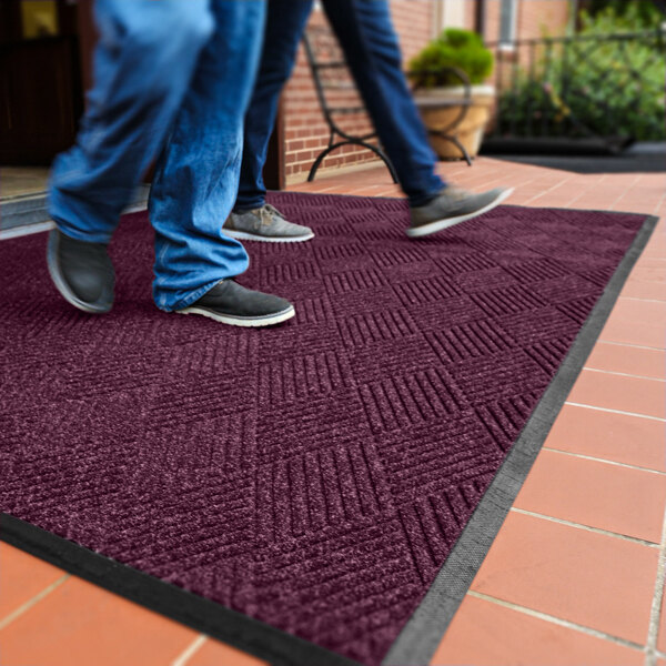 Two people are walking on a large, textured, burgundy mat with a black border placed on a tiled outdoor surface.