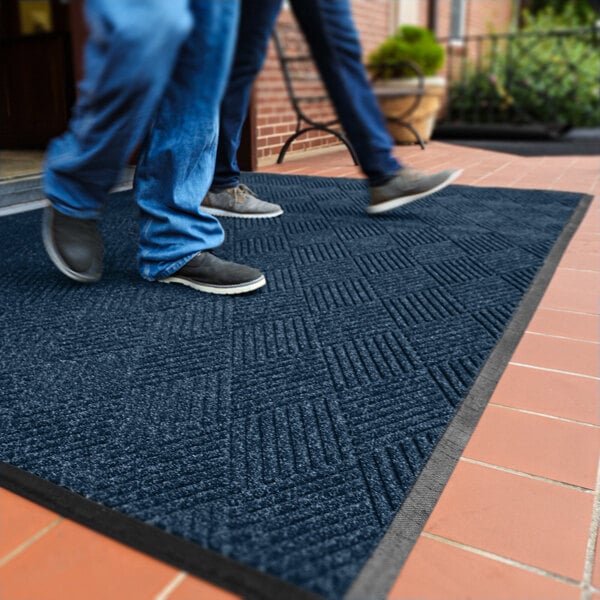 Two people are walking on a dark blue textured mat with a diamond pattern, placed on a tiled outdoor surface.