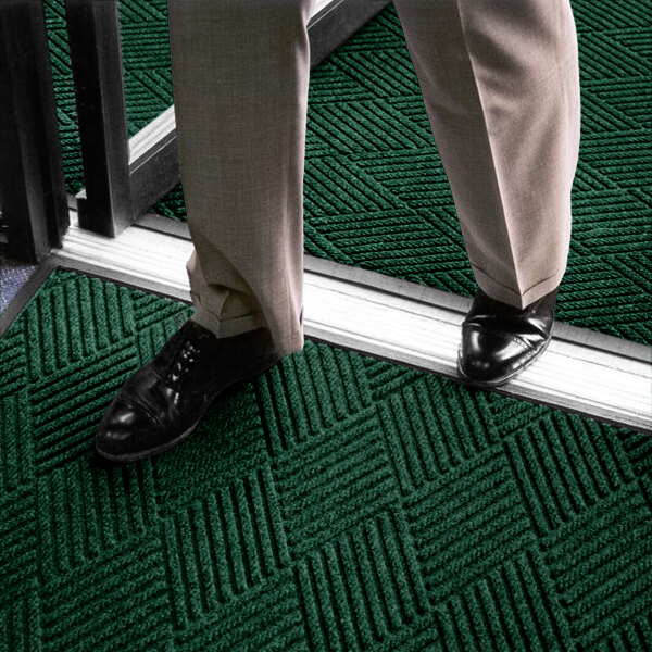 A person wearing dress pants and black shoes is standing on a dark green textured mat with a geometric pattern near a doorway.