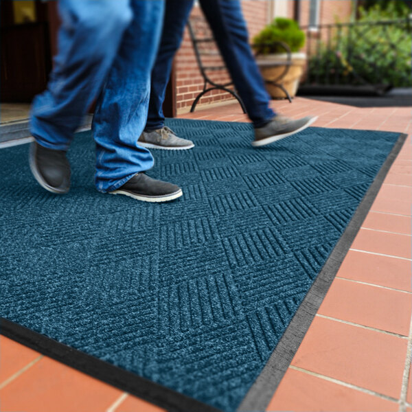 Two people are walking on a large blue textured mat with a diamond pattern, placed on a tiled outdoor surface near a building entrance.