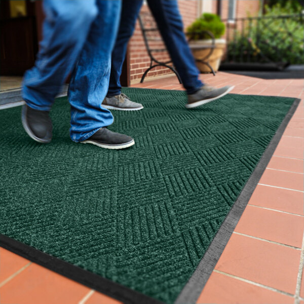 Two people are walking on a dark green textured mat with a diamond pattern and a black border, placed on a tiled floor near an entrance.