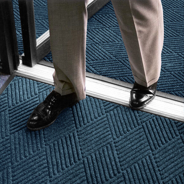 A person wearing dress pants and black shoes is standing on a blue textured mat with a geometric pattern near a doorway.
