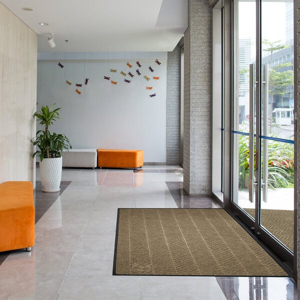 A modern lobby with large glass doors, a brown rectangular floor mat near the entrance, orange and white benches, a potted plant, and colorful wall decorations shaped like birds.