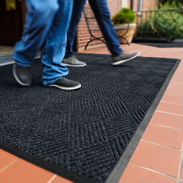 Two people are walking on a large, dark-colored textured mat placed on a tiled outdoor surface near a building entrance.